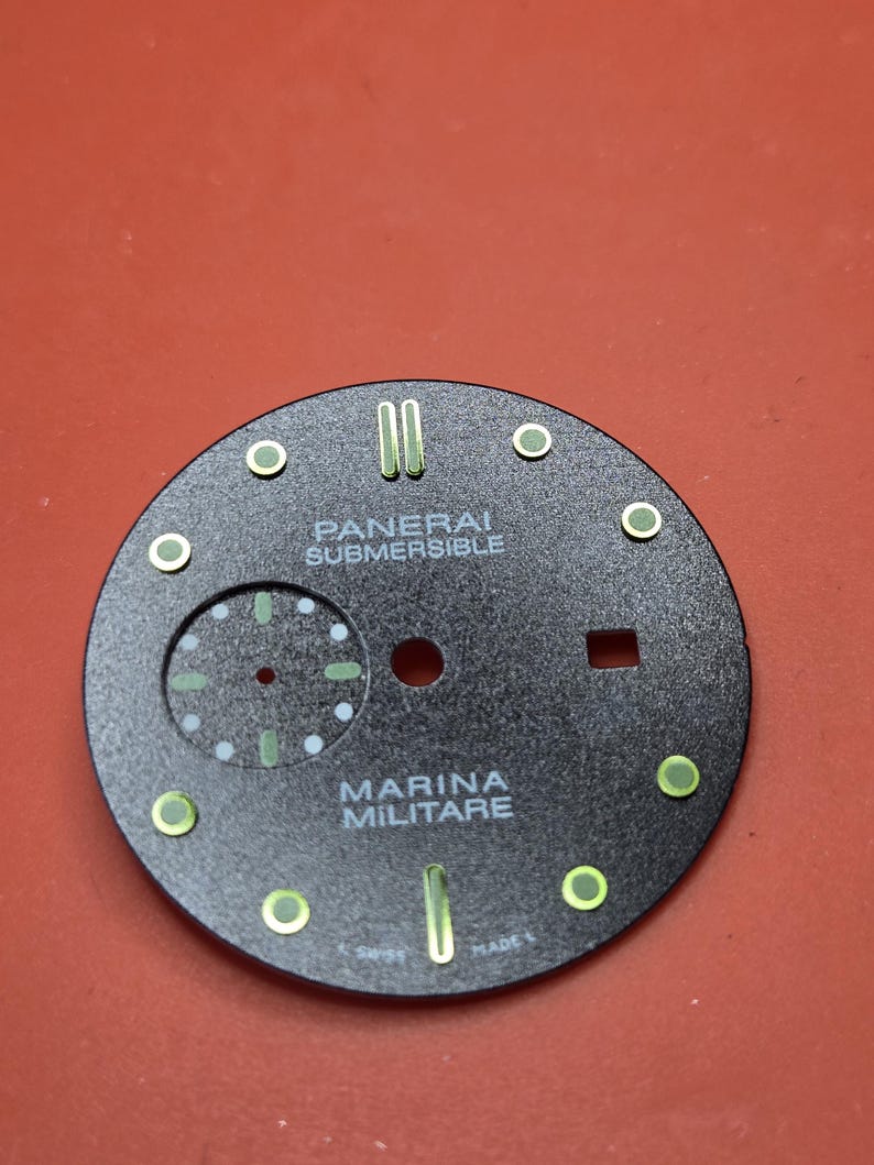 Pode incluir: Um mostrador de rel&oacute;gio preto com as palavras brancas &laquo;PANERAI SUBMERSIBLE&raquo; e &laquo;MARINA MILITARE&raquo;. Possui marcadores verdes e brancos, um pequeno mostrador de segundos e a inscri&ccedil;&atilde;o &laquo;SWISS MADE&raquo;.