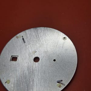 Pode incluir: Um mostrador de rel&oacute;gio prateado com acabamento em metal escovado. O mostrador tem uma abertura quadrada, um furo central e v&aacute;rios furos e pinos menores para fixar os ponteiros e componentes do rel&oacute;gio. O fundo &eacute; de cor vermelha s&oacute;lida.