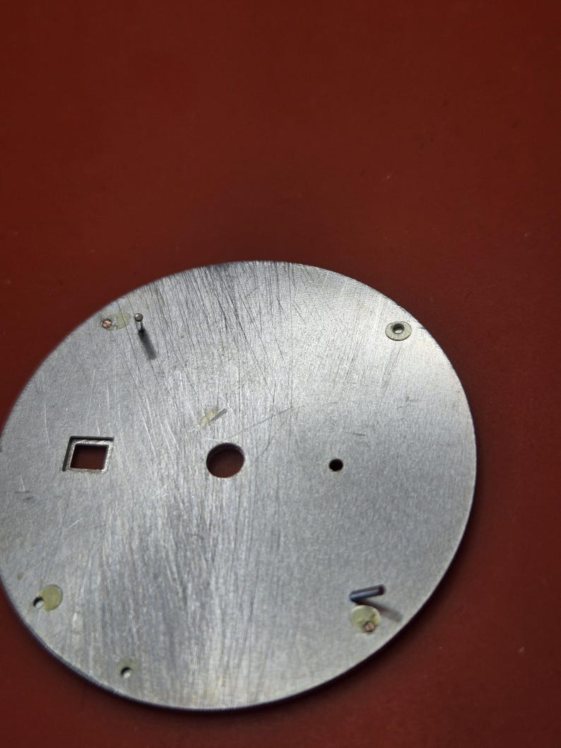 Pode incluir: Um mostrador de rel&oacute;gio prateado com acabamento em metal escovado. O mostrador tem uma abertura quadrada, um furo central e v&aacute;rios furos e pinos menores para fixar os ponteiros e componentes do rel&oacute;gio. O fundo &eacute; de cor vermelha s&oacute;lida.