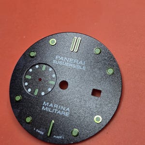 Pode incluir: Um mostrador de rel&oacute;gio preto com o texto branco "PANERAI SUBMERSIBLE" e "MARINA MILITARE". O mostrador tem marcadores circulares e retangulares verdes e ponteiros verdes. O mostrador do rel&oacute;gio &eacute; redondo e tem um pequeno subdial.