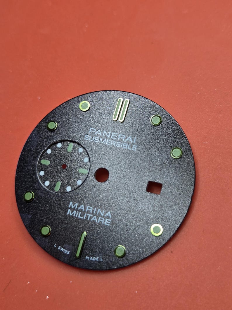 Pode incluir: Um mostrador de rel&oacute;gio preto com o texto branco "PANERAI SUBMERSIBLE" e "MARINA MILITARE". O mostrador tem marcadores circulares e retangulares verdes e ponteiros verdes. O mostrador do rel&oacute;gio &eacute; redondo e tem um pequeno subdial.