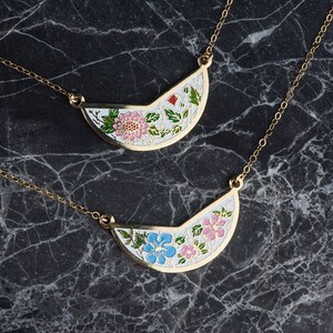 May include: Two gold necklaces with delicate chains and half-circle pendants. The pendants are white with floral designs, one with pink flowers and the other with blue flowers.
