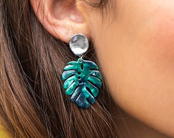 May include: A woman wearing a yellow suede jacket with a silver necklace and a pair of green and blue leaf-shaped earrings.