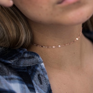 May include: A close-up of a woman wearing a rose gold chain choker necklace. The necklace is delicate and features a simple design. The woman is wearing a blue and white plaid shirt.