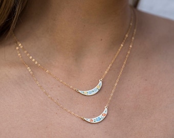 May include: Two gold-colored necklaces with crescent-shaped pendants. Each pendant is white with a floral design in blue, orange, and yellow. The necklaces are layered, with one pendant slightly above the other.