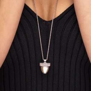 May include: A silver chain necklace with an acorn-shaped pendant. The pendant has a detailed design and a smooth, shiny finish.