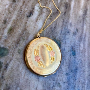 May include: A gold locket necklace with a floral design. The locket has a round shape and is decorated with a delicate floral pattern in shades of pink and gold. The necklace is made of a thin, gold chain.