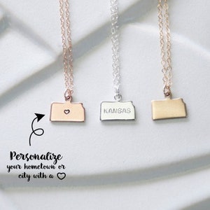 May include: Three delicate necklaces with state-shaped pendants. The pendant on the left is a rose gold outline of a state with a small heart in the center. The middle pendant is silver with the word "KANSAS" engraved on it. The pendant on the right is gold and is a solid outline of a state.  The text "Personalize your hometown or city with a heart" is below the necklaces.