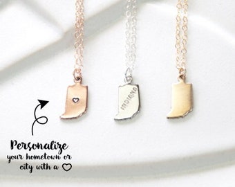 May include: Three gold, silver, and rose gold necklaces with state-shaped pendants. The silver pendant has the word "Indiana" engraved on it. The text "Personalize your hometown or city with a heart" is printed below the necklaces.