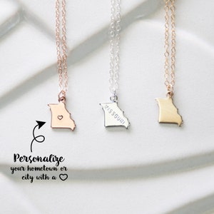 May include: Three gold, silver, and rose gold necklaces with a state of Missouri pendant. The silver pendant has the word "MISSOURI" engraved on it. The rose gold pendant has a small heart in the center. The text "Personalize your hometown or city with a heart" is below the necklaces.