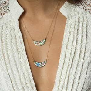 May include: Two gold necklaces with floral pendants. The pendants are shaped like crescent moons and feature colorful floral designs.