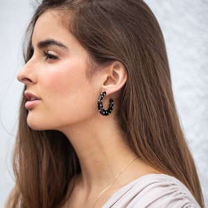 May include: A woman wearing a black hoop earring with gold accents. The earring is made of a smooth, black material and has a dotted pattern of gold studs.