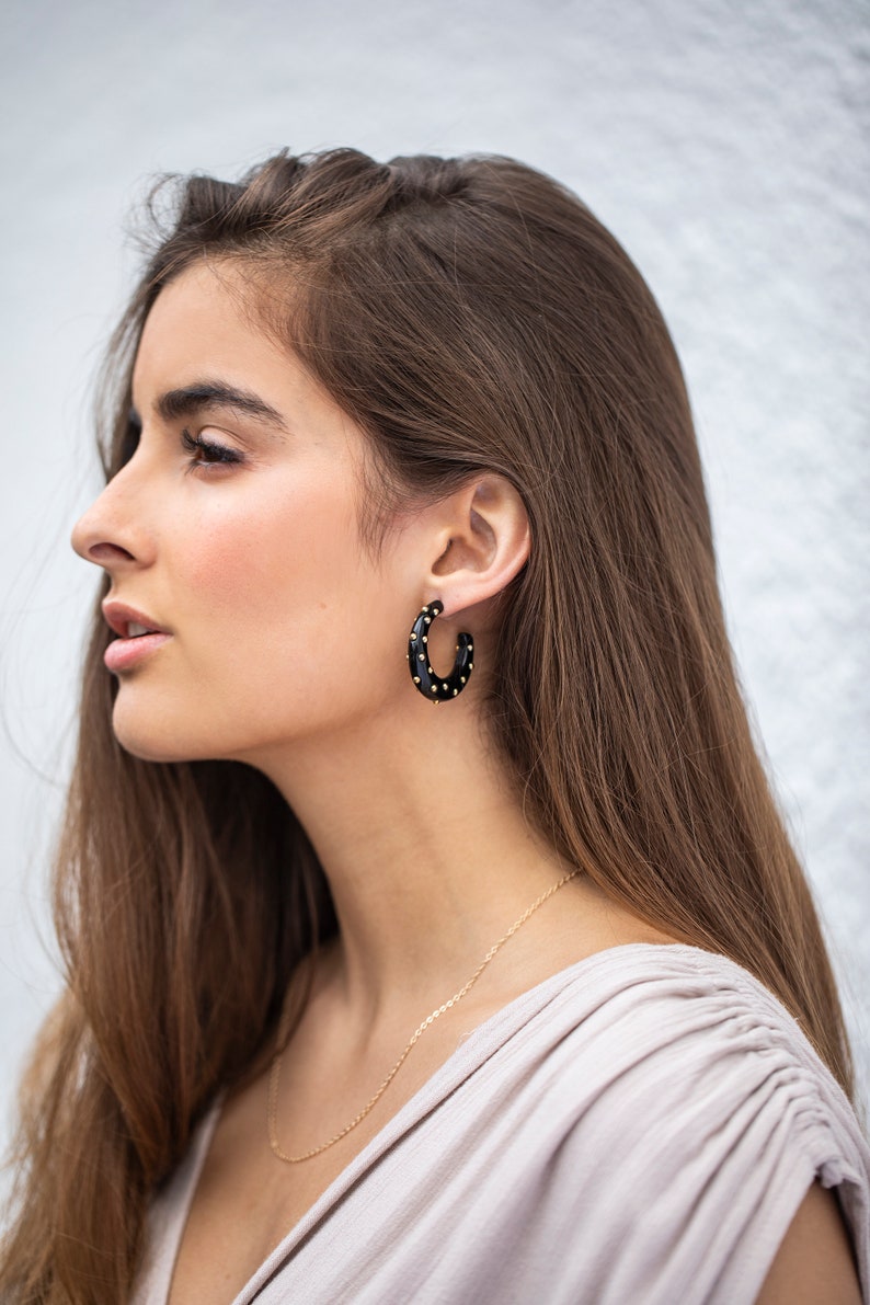 May include: A woman wearing a black hoop earring with gold accents. The earring is made of a smooth, black material and has a dotted pattern of gold studs.