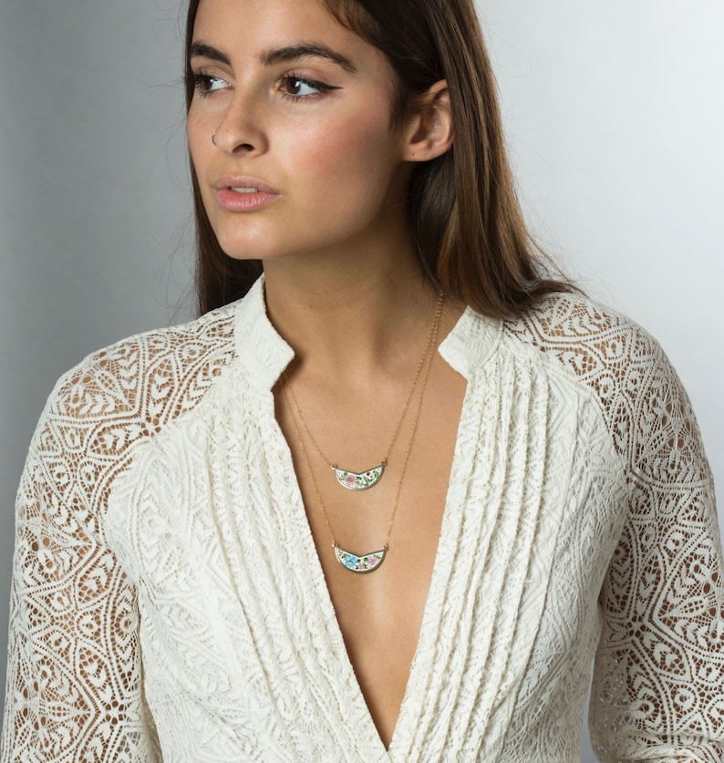May include: A woman wearing a white lace top with a gold chain necklace featuring two floral patterned pendants.