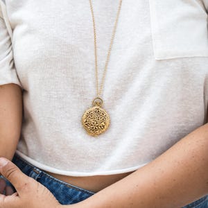 May include: A gold-toned circular pendant necklace featuring an intricate, embossed floral and vine pattern on its surface. The pendant hangs from a delicate gold-toned chain, resting against a plain white t-shirt and blue denim fabric.