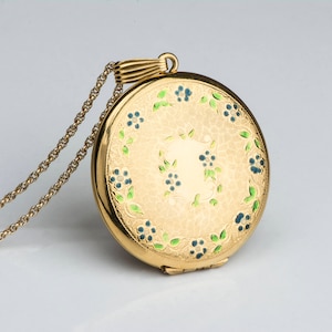 May include: A gold-colored locket necklace with a delicate chain. The round locket features a floral design with blue and green accents. The locket is closed and has a textured surface.