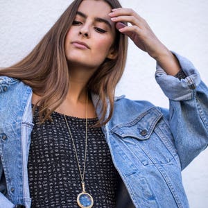 Puede incluir: Una mujer con un vestido de estampado blanco y negro con un collar de cadena dorada y un colgante redondo. Lleva una chaqueta vaquera azul claro y tiene la mano en la cabeza.