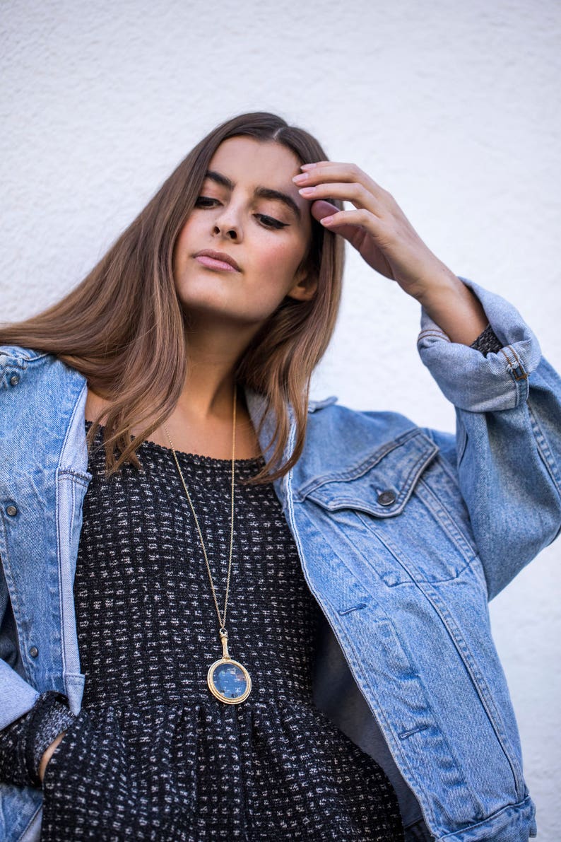 Puede incluir: Una mujer con un vestido de estampado blanco y negro con un collar de cadena dorada y un colgante redondo. Lleva una chaqueta vaquera azul claro y tiene la mano en la cabeza.