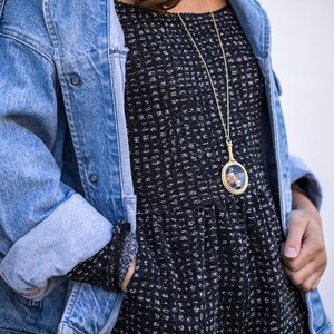 Puede incluir: Una mujer lleva una chaqueta vaquera azul sobre un vestido estampado en blanco y negro. Lleva un collar de cadena dorada con un colgante redondo.
