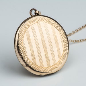 May include: A gold locket necklace with a round, engraved pendant featuring a striped pattern. The pendant has a decorative border.