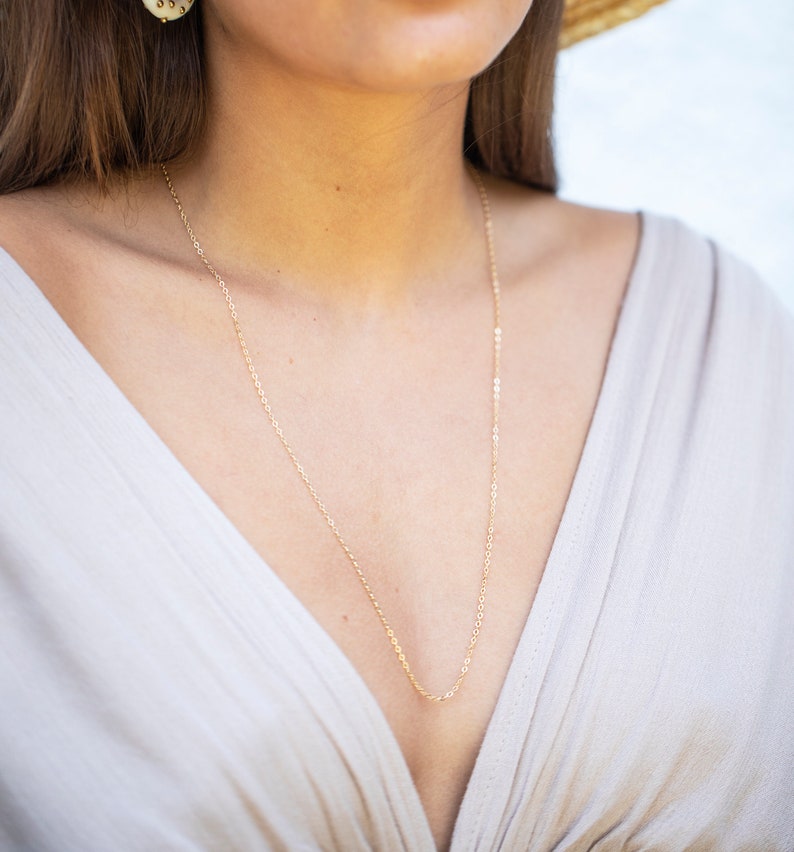 May include: A close-up of a gold chain necklace worn by a person with a low-cut neckline. The chain is thin and delicate, and it hangs down to the person's chest.