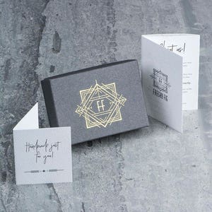 Puede incluir: Una caja gris con un logotipo dorado que presenta las letras "FF" dentro de un dise&ntilde;o geom&eacute;trico. La caja est&aacute; sobre una superficie gris. Una tarjeta blanca con el texto "Handmade just for you!" est&aacute; en primer plano. Una tarjeta blanca con el texto "Freshy Fig" est&aacute; en el fondo.