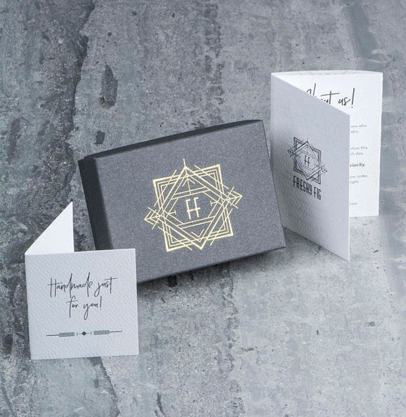 Puede incluir: Una caja gris con un logotipo dorado que presenta las letras "FF" dentro de un dise&ntilde;o geom&eacute;trico. La caja est&aacute; sobre una superficie gris. Una tarjeta blanca con el texto "Handmade just for you!" est&aacute; en primer plano. Una tarjeta blanca con el texto "Freshy Fig" est&aacute; en el fondo.