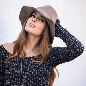 Op de afbeelding: Een vrouw draagt een zwart-wit gepatroneerde trui met een bruine fedora-hoed. Ze draagt een zilveren ketting met een ronde hanger.