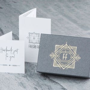 May include: A gray box with a gold geometric design and the letters "FF" in the center. The box is sitting on a gray surface. The text "Handmade just for you!" is on a white card.
