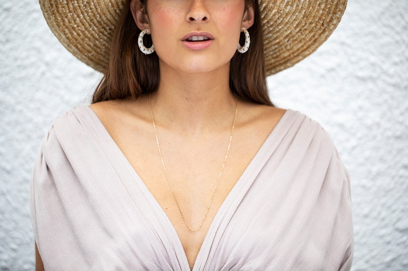 May include: A woman wearing a straw hat with a white top and a gold chain necklace. She is wearing white hoop earrings with gold accents.
