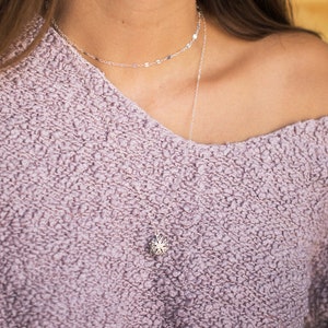 Puede incluir: Un collar de cadena de plata con un delicado colgante con un diseño de estrella. El collar está superpuesto a una segunda cadena más fina con eslabones ovalados.