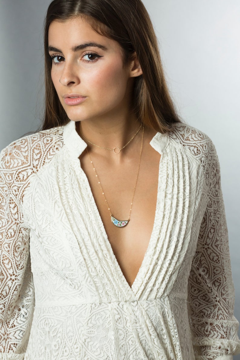 May include: A woman wearing a white lace dress with a gold necklace featuring a crescent moon pendant with a colorful mosaic design.