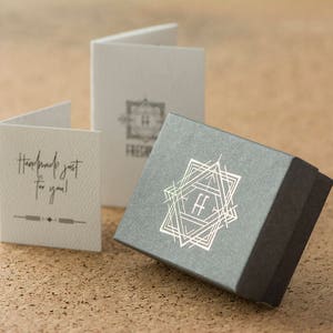 May include: A gray gift box with a silver geometric design and the letters "FF" in the center. The box is sitting on a cork surface. There are two white cards in the background, one with the text "Handmade just for you!" and the other with a geometric design and the text "Freya & Finn".