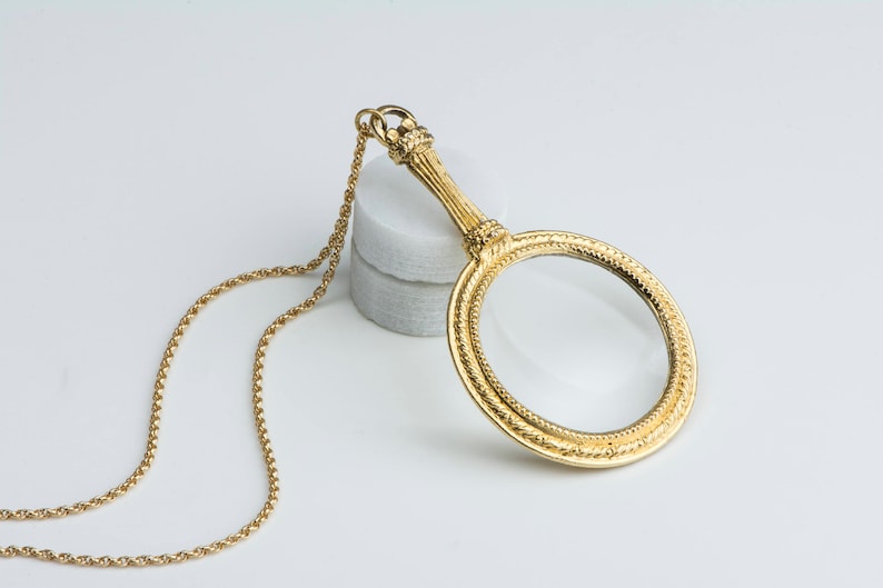 Puede incluir: Un collar de cadena dorada con un colgante de lupa. La lupa tiene un marco redondo adornado y un mango dorado.