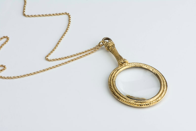 Puede incluir: Un collar de cadena dorada con un colgante de lupa. La lupa tiene un marco dorado y una lente transparente.
