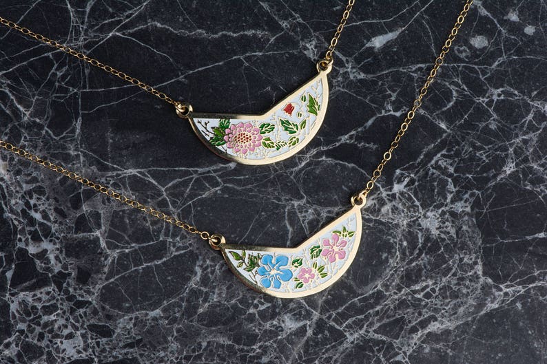 May include: Two gold necklaces with delicate chains and half-circle pendants. The pendants are white with floral designs, one with pink flowers and the other with blue flowers.