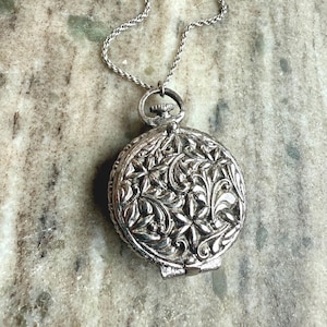 May include: A silver necklace with a round pendant featuring an intricate floral design. The pendant has a hinged opening.