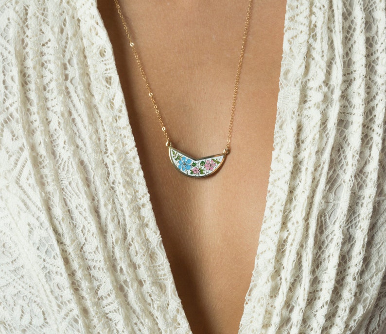 May include: A gold necklace with a crescent moon pendant featuring a floral design in blue, pink, and green enamel.