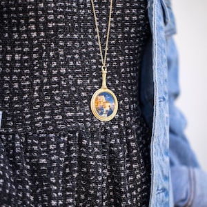 Puede incluir: Un vestido con estampado en blanco y negro con un collar de cadena dorada que presenta un colgante redondo con una imagen colorida en su interior.