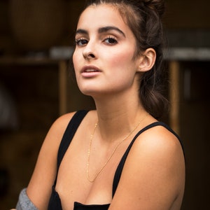 May include: A woman with brown hair pulled back in a bun wears a black tank top and a gray cardigan. She is wearing a gold chain necklace.