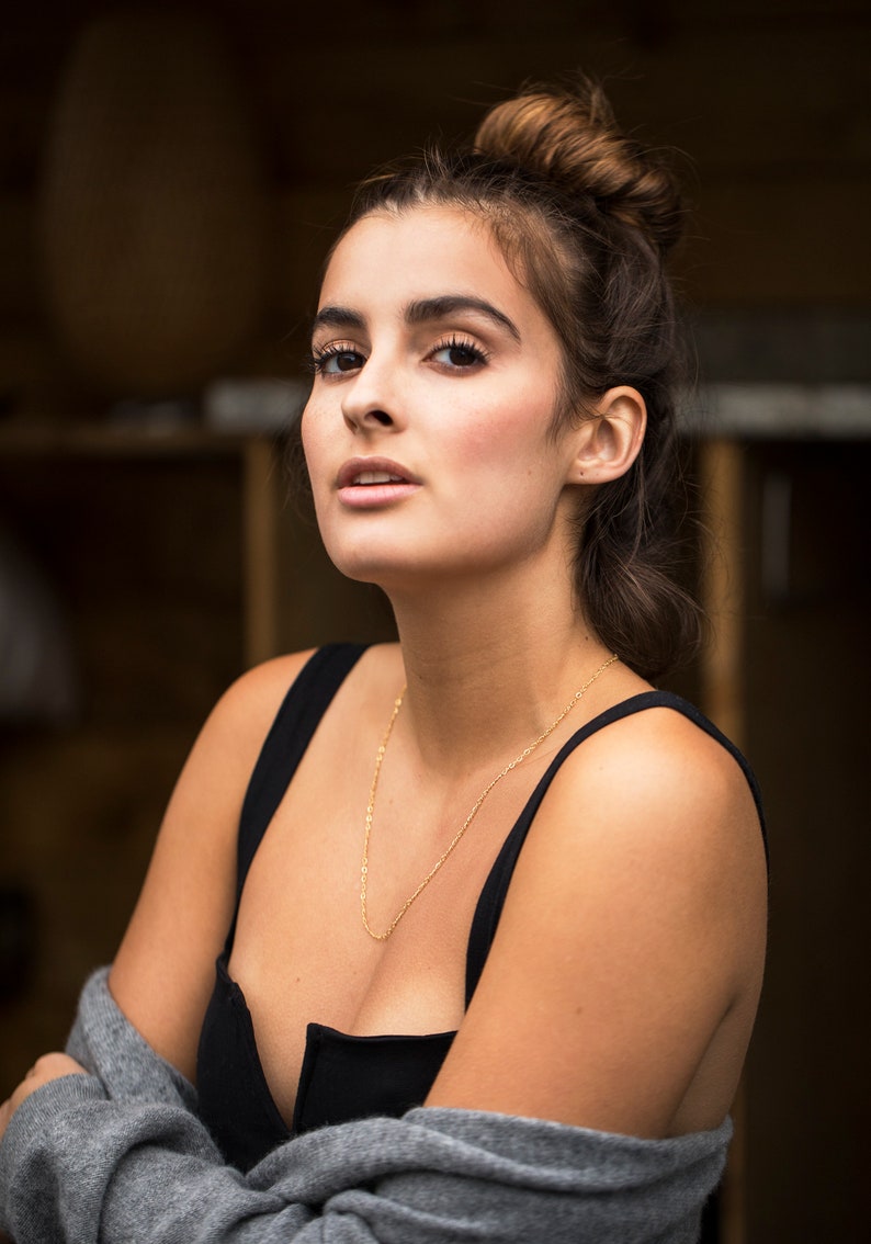 May include: A woman with brown hair pulled back in a bun wears a black tank top and a gray cardigan. She is wearing a gold chain necklace.