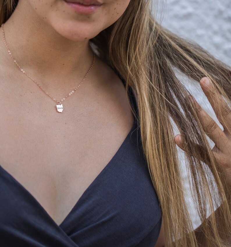 May include: A close-up of a woman wearing a black top and a delicate gold necklace with a small pendant that says "OHIO".