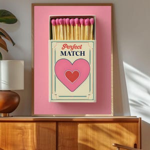 Puede incluir: Impresión enmarcada de un diseño de caja de cerillas con las palabras "Perfect Match" y un corazón rosa. La caja contiene cerillas de madera con cabezas rosas, sobre un fondo rosa. El marco es de color marrón claro.