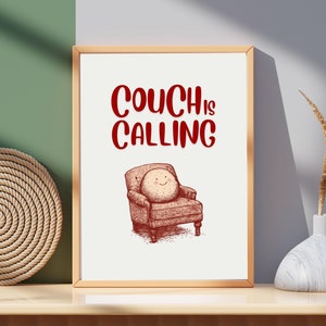 Puede incluir: Una impresión enmarcada con el texto "Couch is Calling" en letras rojas. La impresión presenta una ilustración de dibujos animados de un sofá con una cara sonriente.