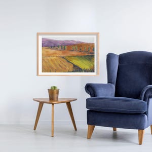 May include: A framed watercolor landscape painting depicting fields, trees, and mountains in shades of yellow, green, orange, and purple. A navy blue armchair and a small wooden side table with a potted cactus are also in the image.