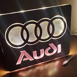 May include: A black light-up sign with the Audi logo and the word "Audi" in pink lettering.