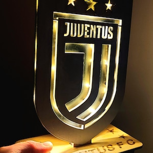 Puede incluir: Un letrero de madera negro con un logotipo de Juventus dorado y tres estrellas doradas. El letrero está iluminado con luces blancas y tiene el texto "Juventus FC" en la base.