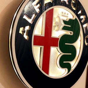 Puede incluir: Un letrero circular de madera con fondo negro y contorno blanco. En el centro del letrero se encuentra el logotipo de Alfa Romeo, que incluye una cruz roja y una serpiente verde. Las palabras "Alfa Romeo" están recortadas en la madera alrededor del logotipo.