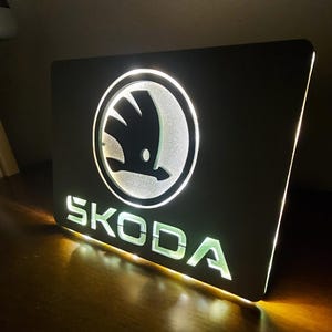 Puede incluir: Un letrero de metal negro con un contorno blanco del logotipo de Skoda y la palabra "SKODA" en letras blancas con un contorno verde.