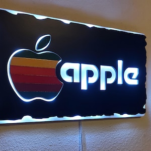 May include: Illuminated sign featuring the Apple logo, a colorful apple with a bite taken out, and the word "apple" in bright white letters. The sign has a black background and a white border with a glowing effect.